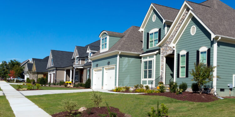 A row of modern suburban houses in a uniform architectural style along a sidewalk and lawn under a clear blue sky
